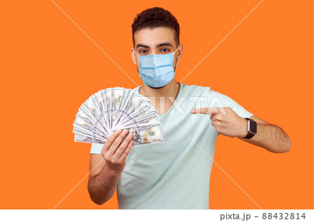 Portrait of lucky rich brunette man with surgical medical mask pointing at dollar banknotes and looking at camera with toothy smile, enjoying wealth. indoor studio shot isolated on orange background Portrait of lucky rich brunette man with surgical medical mask pointing at dollar banknotes and looking at camera with toothy smile, enjoying wealth. indoor studio shot isolated on orange background 88432814