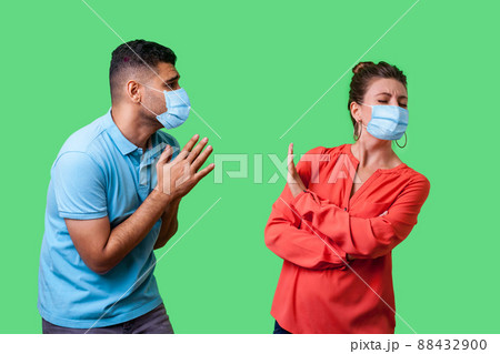 Portrait of couple with surgical medical mask standing together, man holding arms in prayer and pleading forgiveness, looking with hope at naughty woman, isolated on green background, indoor shot Portrait of couple with surgical medical mask standing together, man holding arms in prayer and pleading forgiveness, looking with hope at naughty woman, isolated on green background, indoor shot 88432900