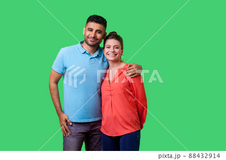 Portrait of young attractive family couple in casual wear standing together, embracing and looking at camera with sincere smile, strong relations. isolated on green background, indoor studio shot Portrait of young attractive family couple in casual wear standing together, embracing and looking at camera with sincere smile, strong relations. isolated on green background, indoor studio shot 88432914