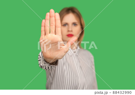 Portrait of serious young woman in classic striped blouse showing stop gesture to camera, warning with raised palm, showing ban, block sign with hand. indoor studio shot isolated on green background Portrait of serious young woman in classic striped blouse showing stop gesture to camera, warning with raised palm, showing ban, block sign with hand. indoor studio shot isolated on green background 88432990