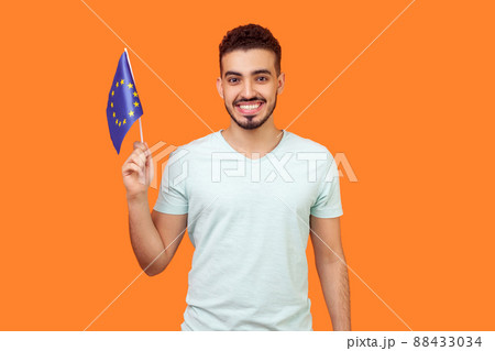 European Union flag. Portrait of happy brunette man with beard in white t-shirt smiling and holding EU flag, looking at camera with toothy smile. indoor studio shot isolated on orange background 88433034