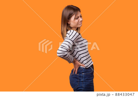 Side view portrait of sick woman with brown hair in long sleeve striped shirt standing with grimace of pain, holding back, suffering strong backache. indoor studio shot isolated on orange background 88433047