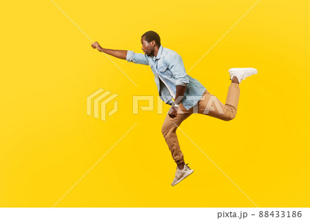 Full length portrait of serious cool handsome man in denim casual shirt jumping in air or flying high with one stretched arm, feeling to be superman. indoor studio shot isolated on yellow background Full length portrait of serious cool handsome man in denim casual shirt jumping in air or flying high with one stretched arm, feeling to be superman. indoor studio shot isolated on yellow background 88433186