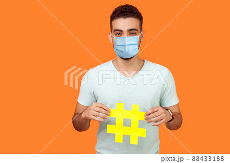 Promotion in social media. Portrait of happy brunette man with surgical medical mask smiling and holding big hashtag sign, sharing viral content. indoor studio shot isolated on orange background Promotion in social media. Portrait of happy brunette man with surgical medical mask smiling and holding big hashtag sign, sharing viral content. indoor studio shot isolated on orange background 88433188