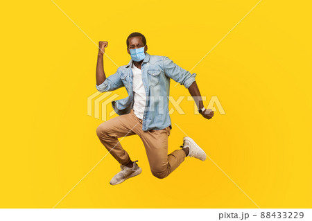 Full length portrait of joyous ecstatic man with medical mask jumping for joy or flying with raised hand, gesturing yes i did it, celebrating success. indoor studio shot isolated on yellow background 88433229