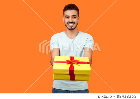 Portrait of happy generous brunette man with beard in white t-shirt smiling and giving gift box to camera, sharing holiday present, charity concept. indoor studio shot isolated on orange background 88433255