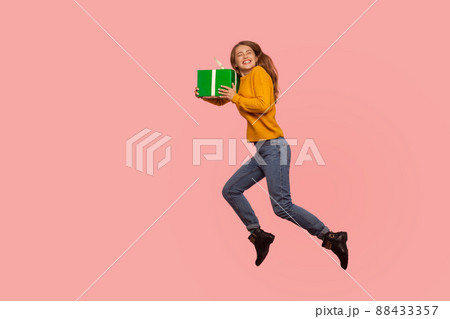 Portrait of extremely happy ginger girl in sweater and denim jumping in air with gift box, full of energy and enthusiasm flying with holiday present. indoor studio shot isolated on pink background Portrait of extremely happy ginger girl in sweater and denim jumping in air with gift box, full of energy and enthusiasm flying with holiday present. indoor studio shot isolated on pink background 88433357