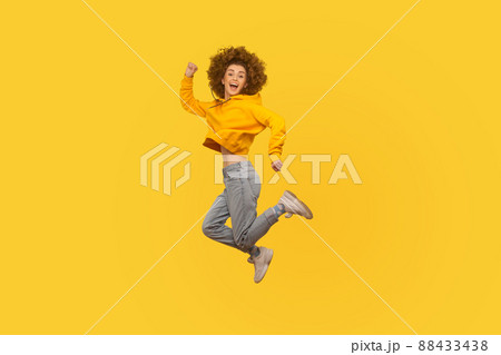 Portrait of enthusiastic joyful curly-haired girl in urban style hoodie and jeans jumping high in air, flying and shouting with amazed happy expression, celebrating success. studio shot isolated Portrait of enthusiastic joyful curly-haired girl in urban style hoodie and jeans jumping high in air, flying and shouting with amazed happy expression, celebrating success. studio shot isolated 88433438