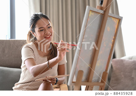 Charming young woman sitting in living room and painting picture with acrylic paint. 88435928