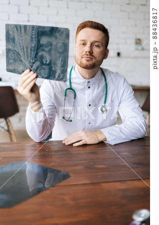 Vertical shot of serious male doctor in white uniform examining brain computerized tomography scan sitting at desk in hospital office, thinking about diagnosis. Concept of medicine and healthcare. 88436817