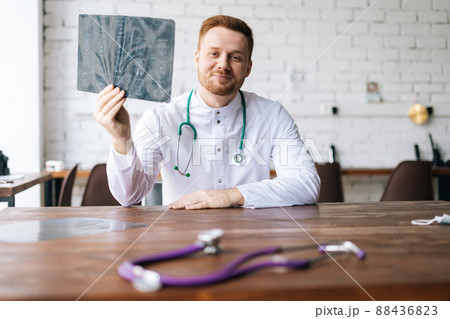 Portrait of happy male doctor in white uniform examining brain computerized tomography scan sitting at desk in hospital office, thinking about diagnosis. Concept of medicine and health care. Portrait of happy male doctor in white uniform examining brain computerized tomography scan sitting at desk in hospital office, thinking about diagnosis. Concept of medicine and health care. 88436823