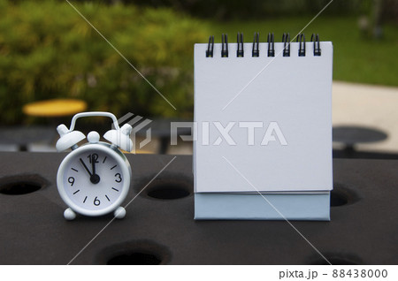 Alarm clock pointing at 11 o'clock with blank notepad. Blurred park background. Copy space concept 88438000