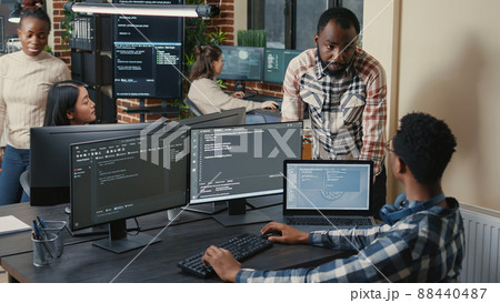 Programer sitting at desk with multiple screens running code talking with colleague about artificial intelligence algorithm. Software developers doing innovative artificial intelligence project. 88440487