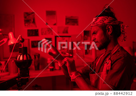 Young photographer of Indian ethnicity standing in red lit darkroom getting ready to develop photos putting on protective gloves 88442912