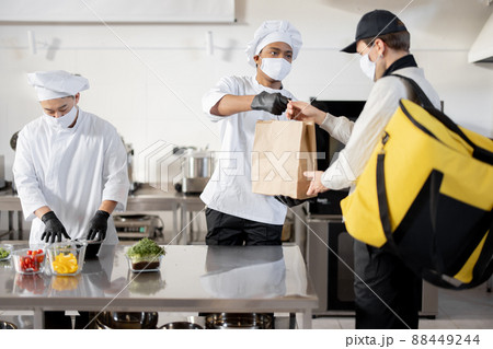 Courier waiting for an order for delivery in the kitchen with chefs preparing takeaway food Courier waiting for an order for delivery in the kitchen with chefs preparing takeaway food 88449244