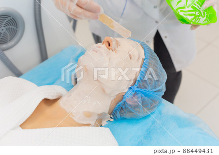The cosmetologist applying an alginate mask to the face of a young lying woman in beauty salon for procedure of cleansing and moisturizing the skin, removing puffiness and lifting effect The cosmetologist applying an alginate mask to the face of a young lying woman in beauty salon for procedure of cleansing and moisturizing the skin, removing puffiness and lifting effect 88449431