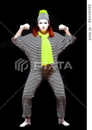 Portrait of female mime artist, isolated on black background. Young woman in striped suit and bright yellow scarf and hat is showing her strength. Symbol energy, power 88449488