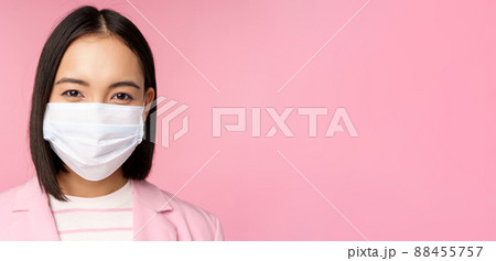 Close up portrait of japanese businesswoman in medical face mask, suit, looking at camera, standing over pink background 88455757