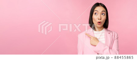 Portrait of saleswoman, korean businesswoman pointing and looking left with surprised, intrigued face expression, posing in suit over pink background 88455865
