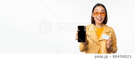 Online shopping and people concept. Stylish asian woman showing mobile phone screen and credit card, smartphone application, standing over white background Online shopping and people concept. Stylish asian woman showing mobile phone screen and credit card, smartphone application, standing over white background 88456021