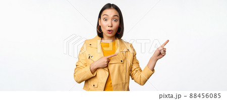 Enthusiastic asian girl pointing fingers right, showing advertisement and smiling, inviting to the store, standing over white background 88456085