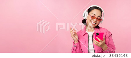 Stylish asian girl dancing with smartphone, listening music in headphones on mobile phone app, smiling and laughing, posing against pink background Stylish asian girl dancing with smartphone, listening music in headphones on mobile phone app, smiling and laughing, posing against pink background 88456148