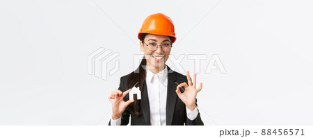 Confident professional female asian architect in helmet and business suit ensure quality construction, showing home maket and okay gesture, recommend company for design and renovation 88456571