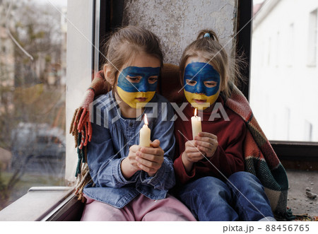 Two little girls with Ukrainian flags painted on their faces are praying for Ukraine, holding lighted candles in their hands, a gesture of faith and hope. Children against war. Two little girls with Ukrainian flags painted on their faces are praying for Ukraine, holding lighted candles in their hands, a gesture of faith and hope. Children against war. 88456765