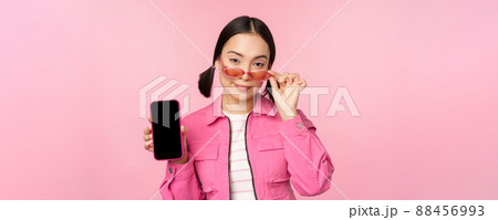 Stylish korean girl, young woman in sunglasses showing smartphone screen, mobile phone app interface or website, standing over pink background Stylish korean girl, young woman in sunglasses showing smartphone screen, mobile phone app interface or website, standing over pink background 88456993