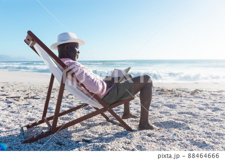 Side view of african american senior man reading book sitting on folding chair at beach 88464666