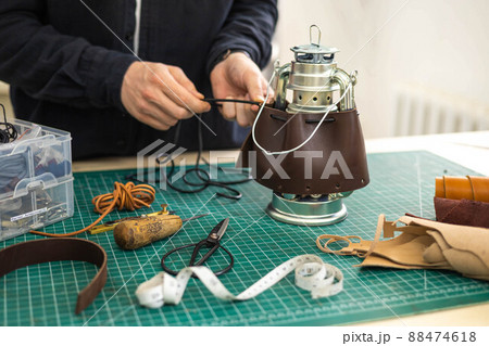 Male tanner working makes a leather case for a camping kerosene lamp. 88474618