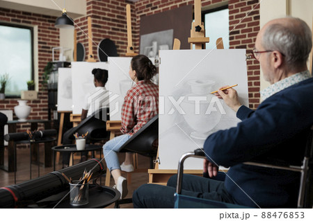 Senior paralyzed man in wheelchair attenting art class drawing vase sketch on white canvas developing sketching skills for personal growth. Diverse group of people creating modern artwork 88476853