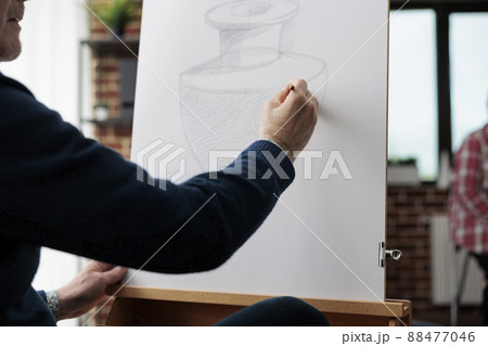 Close up of painter senior man drawing vase sketch on white canvas using illustration pencil during art class in creative workshop. Artist sketching artwork developing graphic technique Close up of painter senior man drawing vase sketch on white canvas using illustration pencil during art class in creative workshop. Artist sketching artwork developing graphic technique 88477046