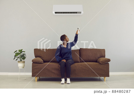 Young african woman switching on air conditioner with remote control at home. AC system concept. 88480287