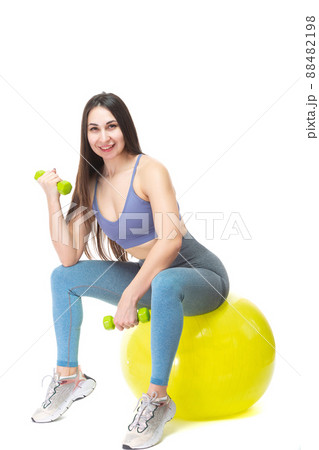 A beautiful, athletic, slender and cheerful woman sits on a yellow fitball and holds light green dumbbells in her hands. Lifestyle concept with sports and gym. Isolated on white background. 88482198