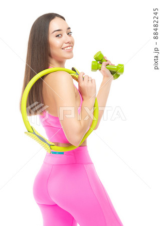 A beautiful, athletic, slim, smiling and cheerful woman in a pink tracksuit is standing with a yellow pilates hoop and green dumbbells. Lifestyle concept with sports and gym. Isolated on white 88482245