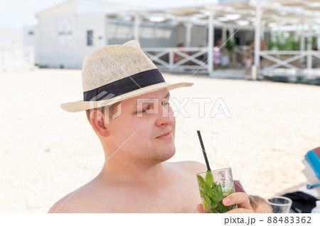 Portrait of young adult caucasian man wearing hat enjoy having fun drinking fresh mojito cocktail and sunbathing on sand beach on hot summer day. Seaside or ocean leisure vacation concept 88483362