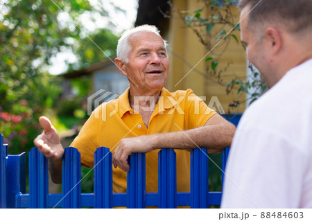 Neighbor conversation. Two smiling men breezily chatting near fence of rural house Neighbor conversation. Two smiling men breezily chatting near fence of rural house 88484603