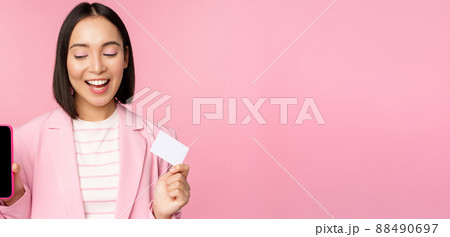 Smiling korean businesswoman in suit, showing mobile phone screen, credit card, showing online banking application interface, pink background Smiling korean businesswoman in suit, showing mobile phone screen, credit card, showing online banking application interface, pink background 88490697