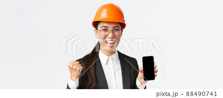Successful winning asian female engineer, architect in safety helmet and suit, showing smartphone screen, fist pump and shouting yes from rejoice, triumphing over achievement, white background Successful winning asian female engineer, architect in safety helmet and suit, showing smartphone screen, fist pump and shouting yes from rejoice, triumphing over achievement, white background 88490741