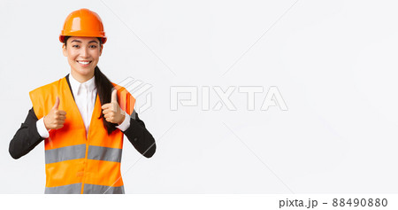 Upbeat pleased asian female architect giving permission, proud with result, standing in safety helmet and reflective jacket, showing thumbs-up in approval, guarantee best construction quality 88490880