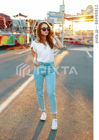 Young hipster woman in stylish T-shirt in trendy sunglasses in blue jeans in sneakers posing in an amusement park on the background of a bright orange summer sunset. Pretty girl enjoying the weekend. Young hipster woman in stylish T-shirt in trendy sunglasses in blue jeans in sneakers posing in an amusement park on the background of a bright orange summer sunset. Pretty girl enjoying the weekend. 88490912