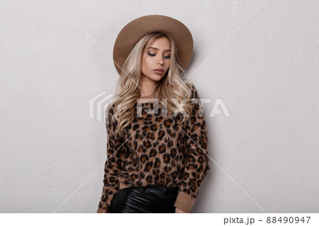 Sensual stylish young woman in a vintage stylish leopard sweater in fashionable black leather pants in a luxurious beige hat posing indoors near a white wall. Cute glamorous girl blonde. 88490947