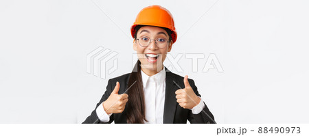 Excited happy female asian engineer, construction manager in safety helmet and business suit showing thumbs-up in rejoice, approve very good work, praise well done job, white background Excited happy female asian engineer, construction manager in safety helmet and business suit showing thumbs-up in rejoice, approve very good work, praise well done job, white background 88490973