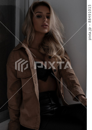 Young gorgeous stylish woman with a beautiful lush breasts in a stylish beautiful lace bra in fashionable leather pants in a beige shirt sitting indoors near the window. Sexy blonde girl. 88491015