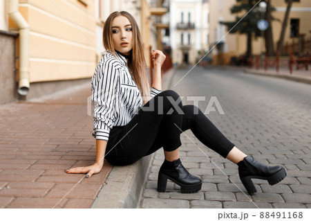 Modern attractive stylish young woman in a fashionable blouse in black jeans in leather shoes is resting sitting on the street near a vintage building.Pretty european girl enjoying summer day outdoors Modern attractive stylish young woman in a fashionable blouse in black jeans in leather shoes is resting sitting on the street near a vintage building.Pretty european girl enjoying summer day outdoors 88491168