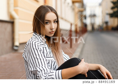 Pretty attractive young woman in a vintage black and white striped blouse in stylish jeans with elegant velvet necklaces sits in the city on a warm spring day. Beautiful girl relaxes outdoors. Pretty attractive young woman in a vintage black and white striped blouse in stylish jeans with elegant velvet necklaces sits in the city on a warm spring day. Beautiful girl relaxes outdoors. 88491169