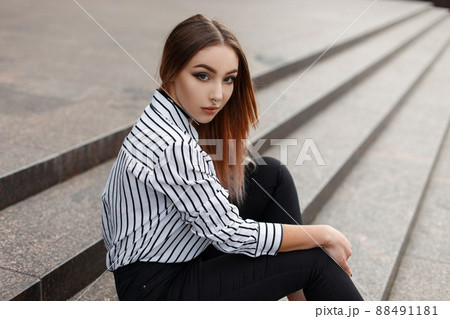 Pretty American young woman in fashionable jeans in a vintage black and white striped blouse with elegant velvet necklaces sits on the vintage steps in the city on a warm spring day. Beautiful girl. Pretty American young woman in fashionable jeans in a vintage black and white striped blouse with elegant velvet necklaces sits on the vintage steps in the city on a warm spring day. Beautiful girl. 88491181