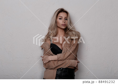Elegant stylish attractive young woman with beautiful magnificent breasts in a lace bra in vintage beige stylish shirt in leather black pants in a room near a white wall. Sexy blonde girl. 88491232