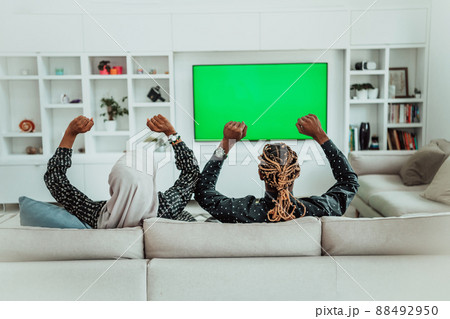 African Couple Sitting On Sofa Watching TV Together Chroma Green Screen Woman Wearing Islamic Hijab Clothes 88492950